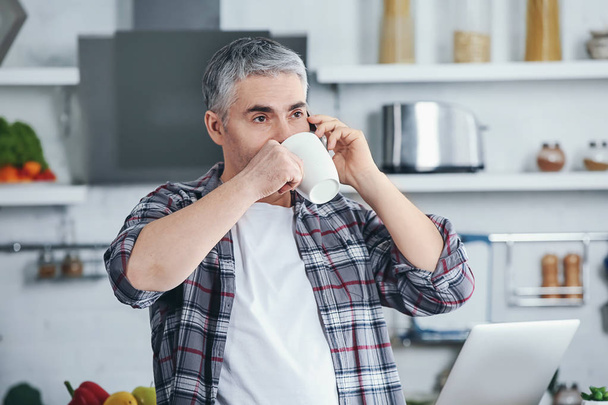 Mature man talking by phone while drinking coffee in kitchen - Foto, Imagen