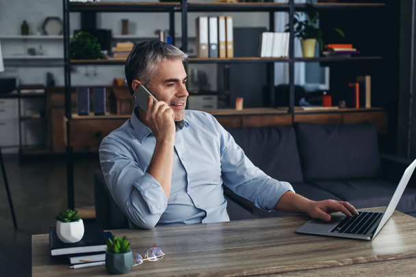 Hombre maduro hablando por teléfono mientras trabaja en el ordenador portátil en la oficina
 - Foto, Imagen