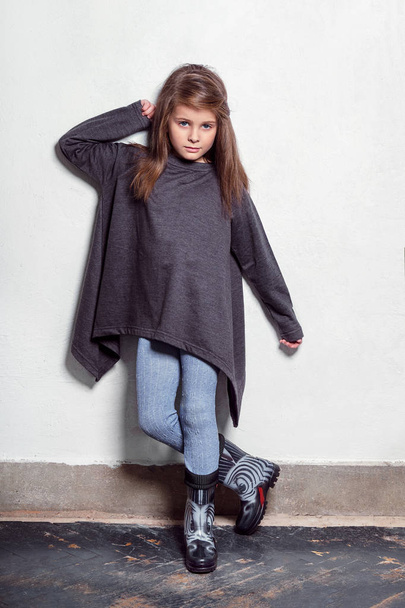 Fashion young model kid posing in a stylish, urban, casual clothes. Little child looks with a serious, hand touching her hair, standing near white wall. childrens leggings, sweater, rubber boots.. - Foto, Bild