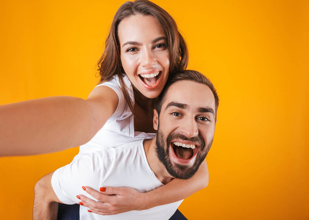 Image of excited couple taking selfie photo while man piggybacki - Foto, afbeelding