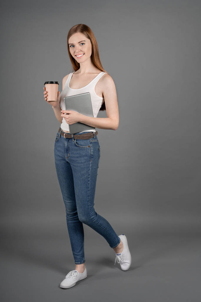 Vertical shot of smiling business woman with the tablet and cup of coffee in her hands looking at camera while standing against grey background - Fotografie, Obrázek