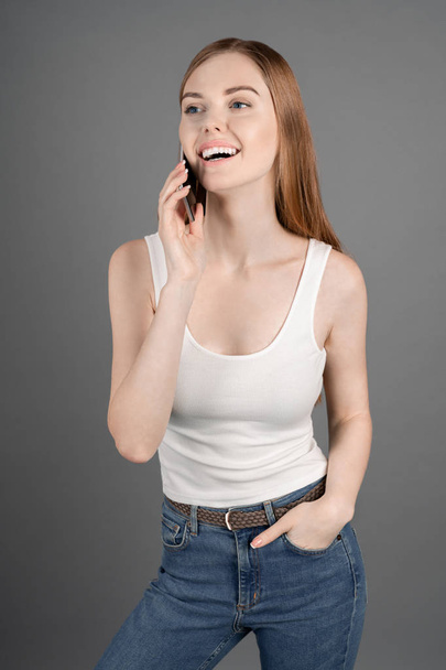 Vertical shot of smiling young woman in casuals talking on mobile phone standing against grey background - Fotografie, Obrázek