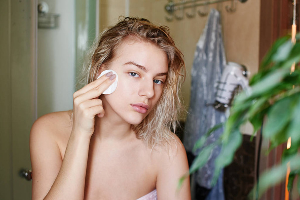 A beautiful girl after shower looks into the camera as in mirror and cleans her face from make up with a cotton pad in the bathroom - Foto, Bild