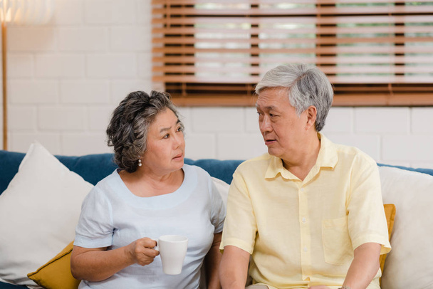 Asian elderly couple drinking warm coffee and talking together in living room at home, couple enjoy love moment while lying on sofa when relaxed at home. Lifestyle senior family at home concept. - Φωτογραφία, εικόνα