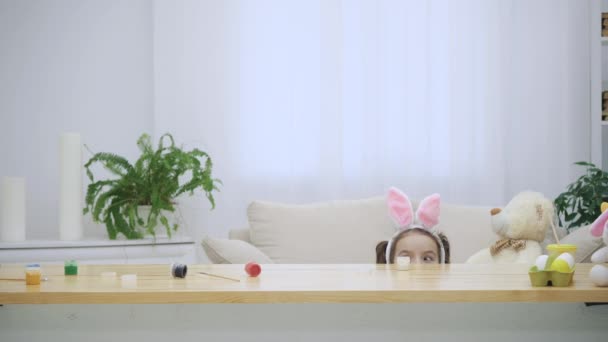 Little playing kid with bunny ears on their heads is hiding under the wooden table, full of Easter decorations. Cute girl is looking out with her funny father. Thery rush in a big hug at the end - Footage, Video