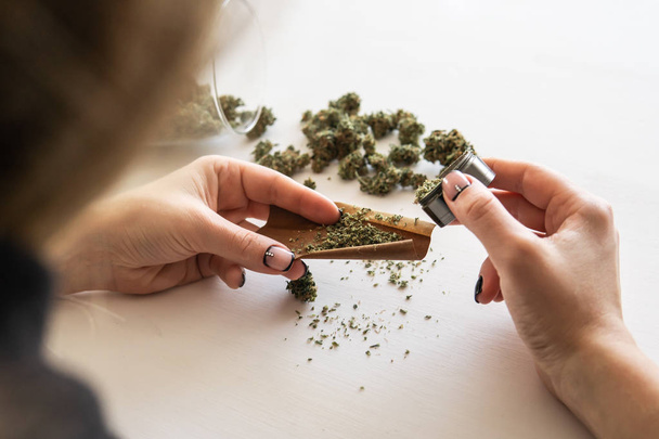 Woman preparing and rolling marijuana cannabis joint. Close up of marijuana blunt with grinder. Cannabis use concept. Woman rolling a marijuana blunt on white background. - Photo, image