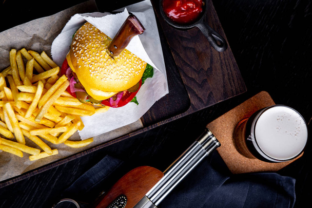 Ремесленный говяжий бургер и картошка фри на столе в ресторане со стаканом пива на тёмном фоне. Современная рамка для ланча
 - Фото, изображение