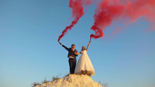 Happy bride and groom waving colored pink smoke against blue sky and laughing. Honeymoon. Romance. Relationship between man and woman - Photo, Image