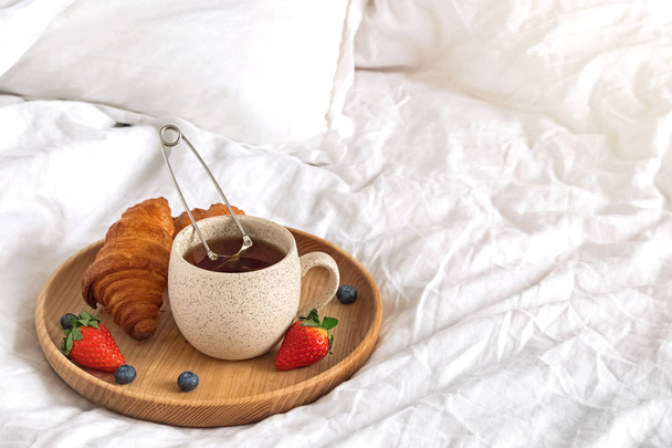 A cup of tea and croissants on the wooden tray standing on the bed with white bedsheets. - Фото, изображение