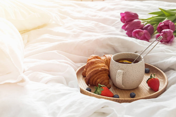 A cup of tea and croissants on the wooden tray standing on the bed with white bedsheets. - Foto, Imagem