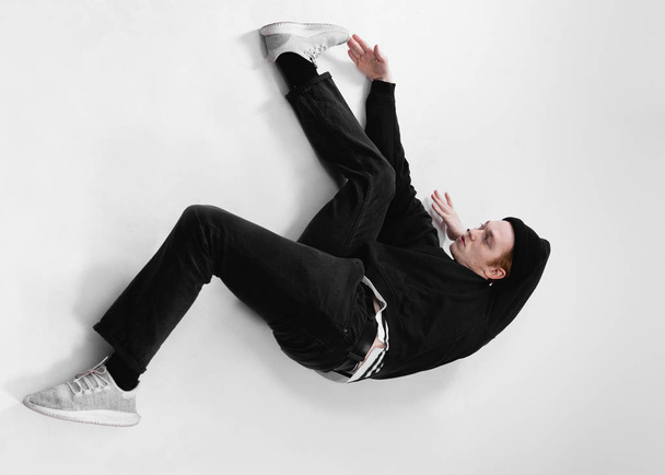 Freestyle dancer dressed in black jeans, sweatshirt, hat and gray sneakers is dancing lying on the floor in the studio on the white background - Photo, Image
