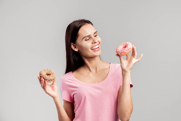 Smiling girl dressed in pink t-shirt holds two mouth-watering donuts in her hands on the white background in the studio - Фото, зображення