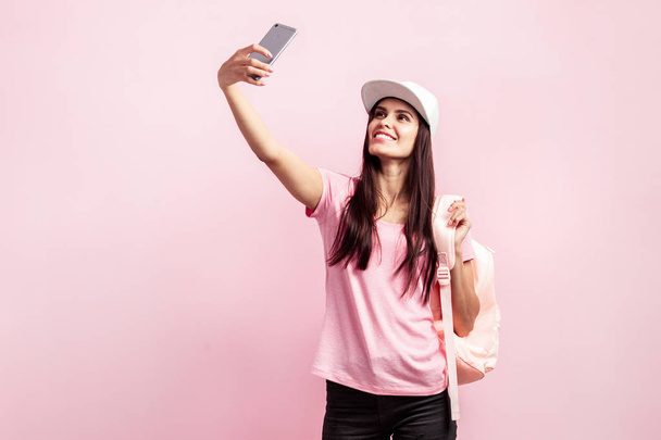 Beautiful girl in white cap and with a backpack on her shoulders dressed in pink t-shirt and black jeans is doing selfie on the pink background in the studio - Fotoğraf, Görsel