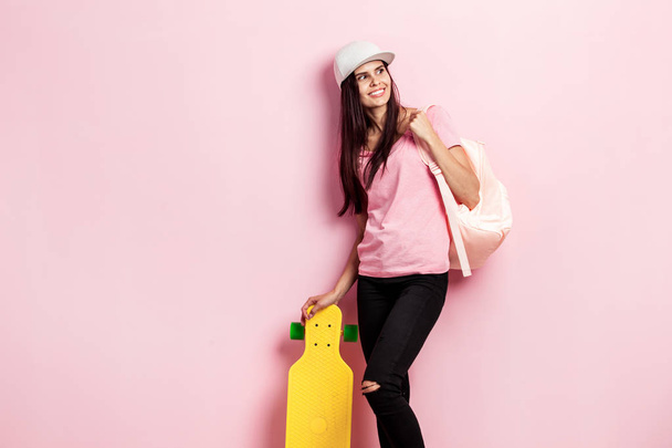 Beautiful girl in white cap and with a backpack on her shoulder dressed in pink t-shirt and black jeans stands with yellow skateboard on the pink background in the studio - Fotó, kép