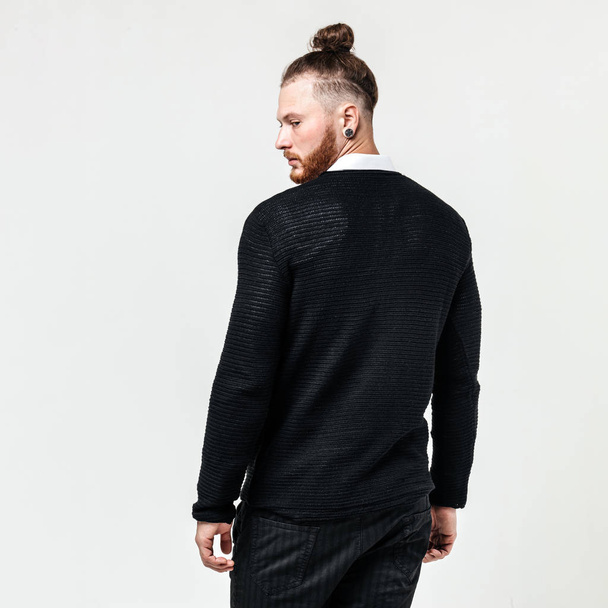 Stylish man with beard and bun hairstyle wearing a black jumper over a white shirt and trousers poses in the studio on the white background - Fotoğraf, Görsel