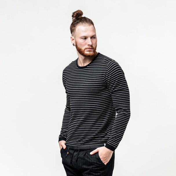 Stylish man with beard and bun hairstyle wearing a black jumper over a white shirt and trousers poses in the studio on the white background - Photo, image