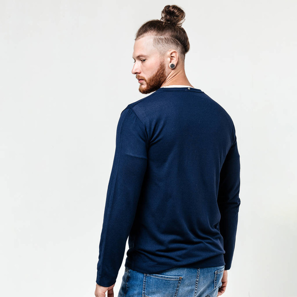 Stylish man with beard and bun hairstyle dressed in blue long sleeve sweater and jeans poses in the studio on the white background - Photo, image