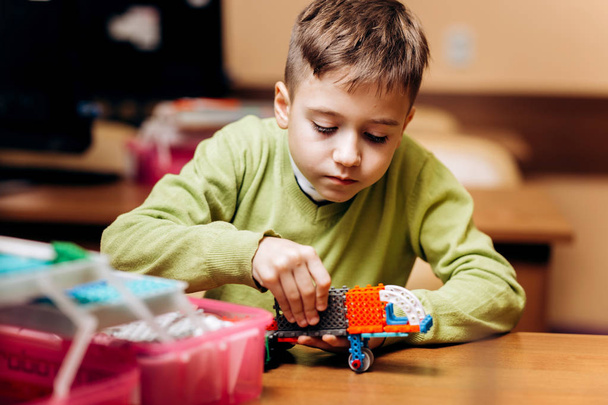 Boy dressed in green sweater sits at the table in the robotics school and makes a robot from the robotic constructor - Foto, immagini