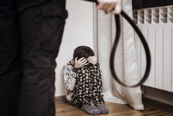 Aggressive parent with belt and frightened child in corner. Domestic violence. Abused child, aggression in the family - Photo, Image