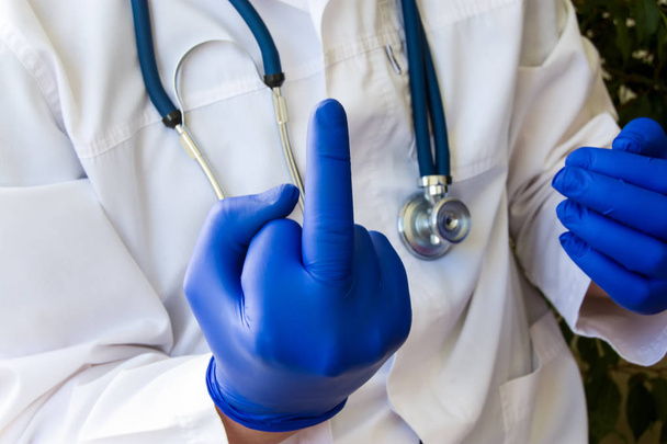 Fuck you sign fingers of hand by doctor or health worker. Doctor showing obscene sign fuck you with raised of middle finger of hand, dressed in blue latex glove in white medical gown on background - Photo, Image
