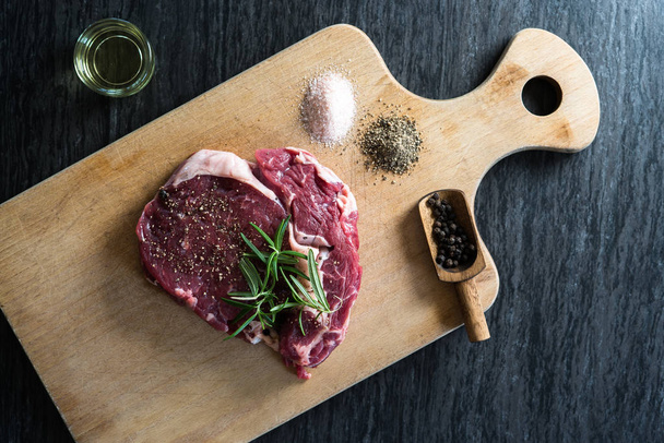 pedaço de carne crua fresca com pimenta, sal e alecrim pronto para cozinhar em tábua de madeira
 - Foto, Imagem