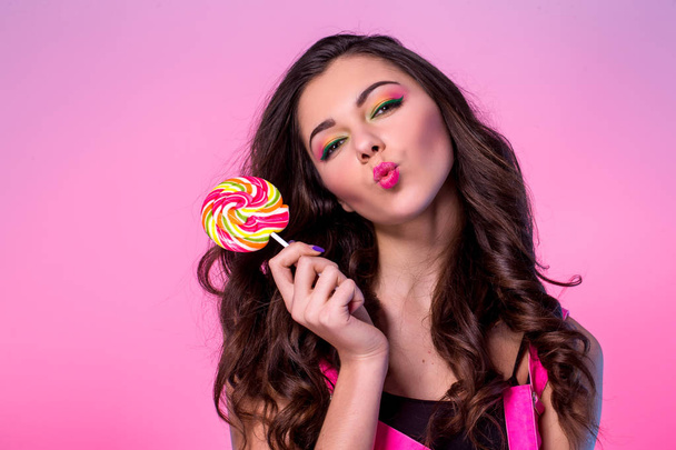 Funny young girl in jumpsuit holding lollipop and posing for camera across pink background - Foto, imagen