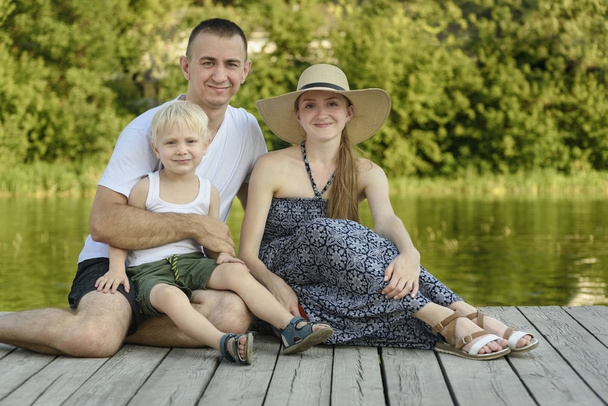 Mladá rodina, otec matku a malá blonďatá syn sedí na řeky molo - Fotografie, Obrázek