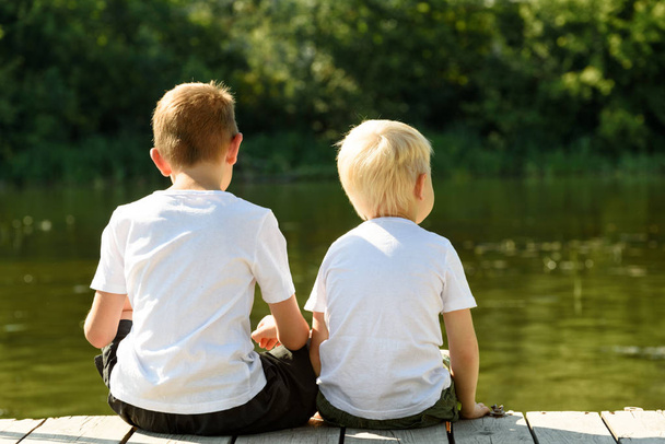 Два маленьких мальчика сидят на пирсе на берегу реки. Понятие дружбы и братства. Вид сзади
 - Фото, изображение
