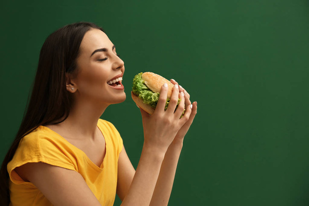 Young woman eating tasty burger on color background. Space for text - Φωτογραφία, εικόνα