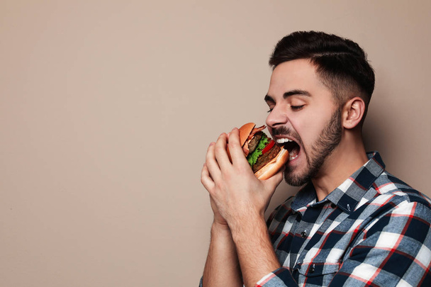 Handsome man eating tasty burger on color background. Space for text - Foto, afbeelding