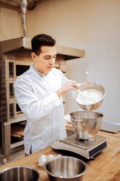 Dark-eyed professional baker adding some flour into bowl - Fotó, kép
