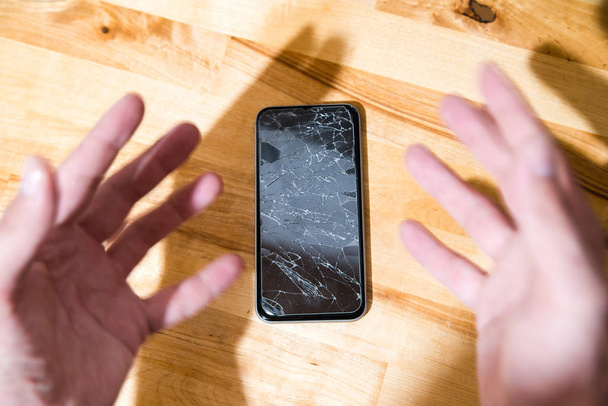 Concept of falling smart phone with broken screen. Top view on wooden desk background. Cracked, shattered lcd touch screen on modern cellphone. Gadget needs repairing - Photo, Image