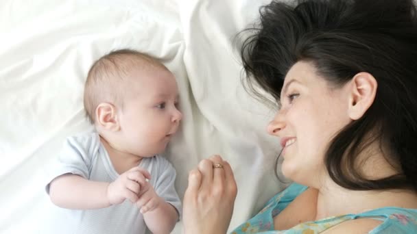 Eine schöne junge Mutter und ein lustiges zwei Monate altes Neugeborenes liegen auf dem Bett und schauen einander an. Mutter streichelt ihr Baby sanft. - Filmmaterial, Video