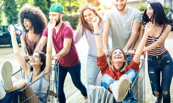 Young multiracial people having fun together with shopping cart - Millenial friends sharing time with trolleys at commercial mall parking - Youth lifestyle concept with focus on girl with hands up - Photo, Image