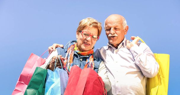 Casal sênior compras em conjunto com a esposa assistindo em sacos de marido - Conceito de idoso com homem maduro e mulher se divertindo no dia ensolarado - Momentos felizes das pessoas aposentadas no filtro vívido contra o céu azul
 - Foto, Imagem