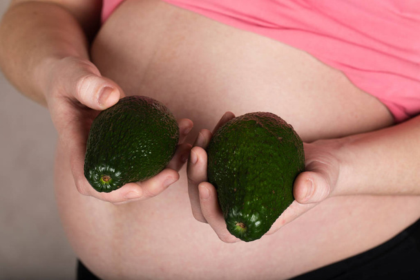 Jonge zwangere vrouw houdt twee avocado's dicht bij haar buik.  - Foto, afbeelding