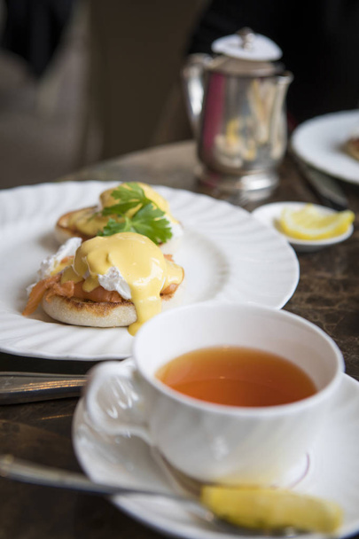 Elegant eggs benedct on a white plate with hollaindaise saice and English tea on top of table. English tea room breakfast. - Фото, зображення