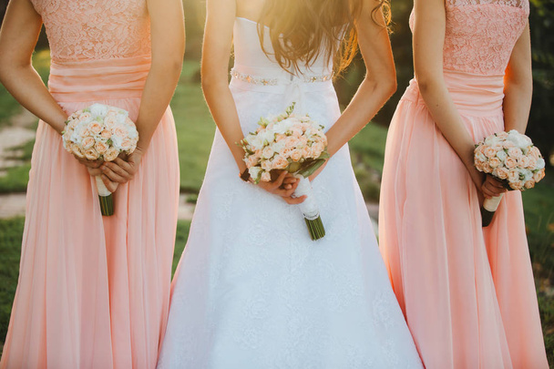 エレガントなドレスを着た花嫁と新婦付添人は、パステルピンクの花と緑のリボンの手の花束を自然に立って保持しています。若いです美しい女の子は屋外で結婚式の花束を保持. - 写真・画像