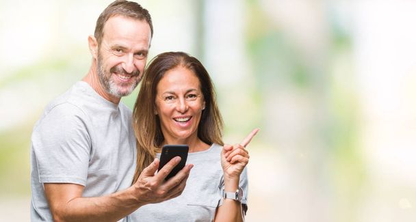 Mediana edad pareja hispana mensaje de texto en el teléfono inteligente ver fondo aislado muy feliz señalando con la mano y el dedo a un lado
 - Foto, Imagen