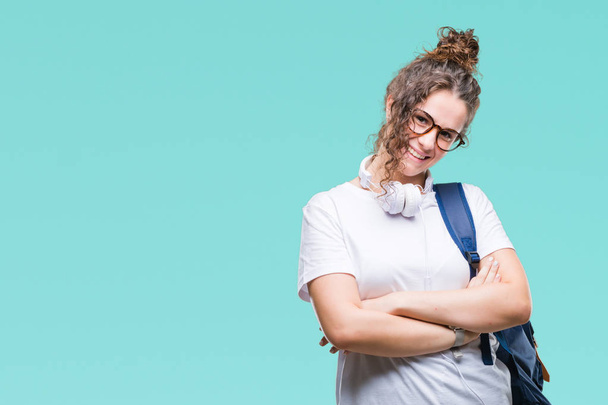 Young brunette student girl wearing backpack and headphones over isolated background happy face smiling with crossed arms looking at the camera. Positive person. - Zdjęcie, obraz