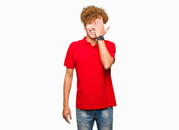 junger gutaussehender Mann mit Afro-Haaren trägt rotes T-Shirt, das ein Auge mit einer Hand bedeckt, mit selbstbewusstem Lächeln im Gesicht und überraschender Emotion. - Foto, Bild