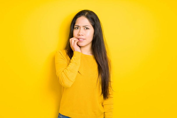 Beautiful brunette woman over yellow isolated background looking stressed and nervous with hands on mouth biting nails. Anxiety problem. - Fotó, kép