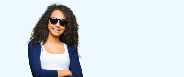Όμορφη κοπέλα με τα σγουρά μαλλιά φορώντας γυαλιά ηλίου μόδας χαρούμενο πρόσωπο χαμογελά με σταυρωμένα χέρια, κοιτάζοντας την κάμερα. Θετικό πρόσωπο. - Φωτογραφία, εικόνα