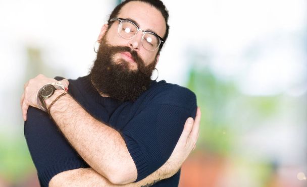 Homem jovem hipster com cabelos longos e barba usando óculos Abraçando-se feliz e positivo, sorrindo confiante. Auto amor e auto cuidado
 - Foto, Imagem