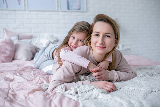 Beautiful young mother and her little daughter are lying together on the bed in the bedroom, playing, hugging and having fun. Maternal care and love. Horizontal photo - Φωτογραφία, εικόνα