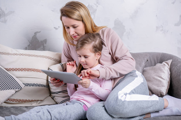 Beautiful young mother and her little daughter are watching movies together and playing on the tablet while sitting on the sofa. Mom hugs daughter. Maternal care and love. Horizontal photo - Фото, зображення