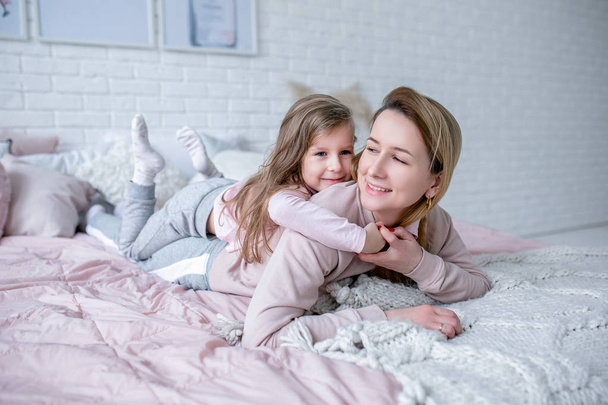 Beautiful young mother and her little daughter are lying together on the bed in the bedroom, playing, hugging and having fun. Maternal care and love. Horizontal photo - Photo, image