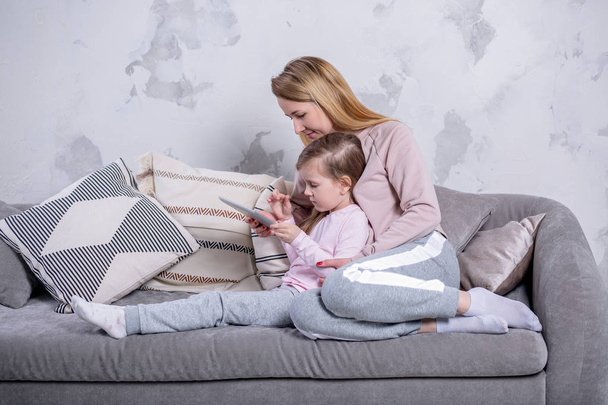 Beautiful young mother and her little daughter are watching movies together and playing on the tablet while sitting on the sofa. Mom hugs daughter. Maternal care and love. Horizontal photo - Φωτογραφία, εικόνα