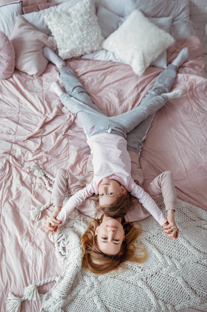 Beautiful young mother and her little daughter are lying together on the bed in the bedroom, playing, hugging and having fun. Maternal care and love. Horizontal photo - Foto, afbeelding