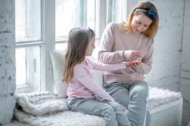 Beautiful young mother and her little daughter are sitting by the window together and painting their nails. Maternal care and love. Horizontal photo - Fotografie, Obrázek